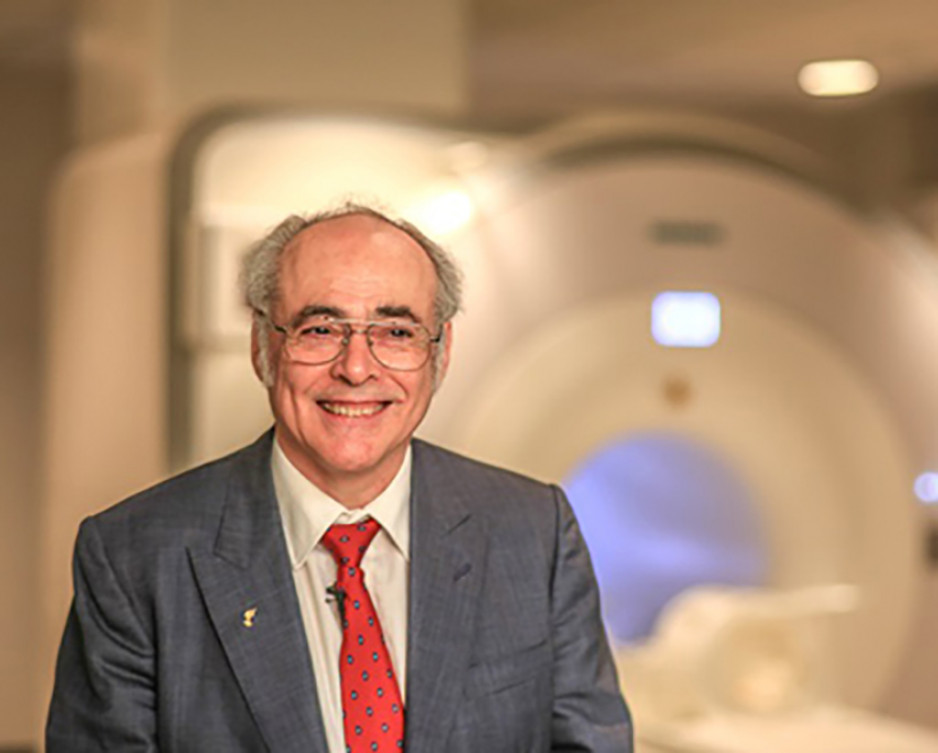 a man with grey hair wearing a grey suit , white shirt and red tie, stands before the blurred out image of an mri machine