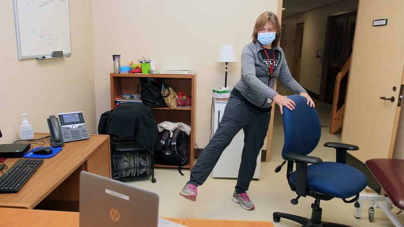 a physiotherapist demonstrates a leg raise exercise while looking at her patient on a laptop montior