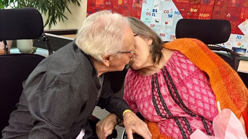 Elderly man and woman are seated beside each other and are are kissing with their faces away from the camera