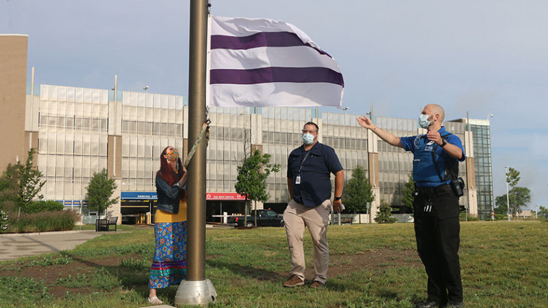 an Indigenous woman raises the Wampum flag at Parkwood Institute, as two flag holders look up at it