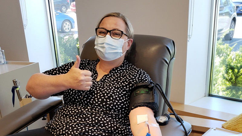  A masked woman in glasses, dress pants and a blouse reclines on a hospital bed with one arm outstretched as she gives blood and the other giving a thumbs up