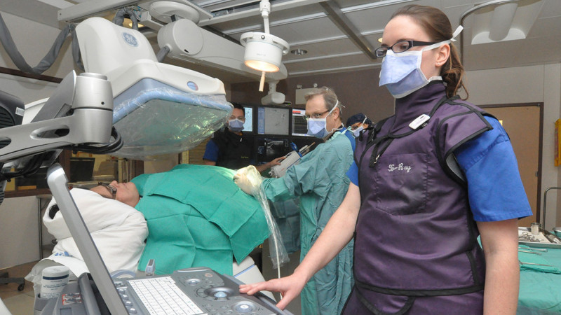 A woman in a purple vest and blue scrubs wearing glasses stands in the foreground of the picture beside a patient on a hospital stretcher, who is under some imaging equipment. Two men monitor the patient at the foot of the stretcher.