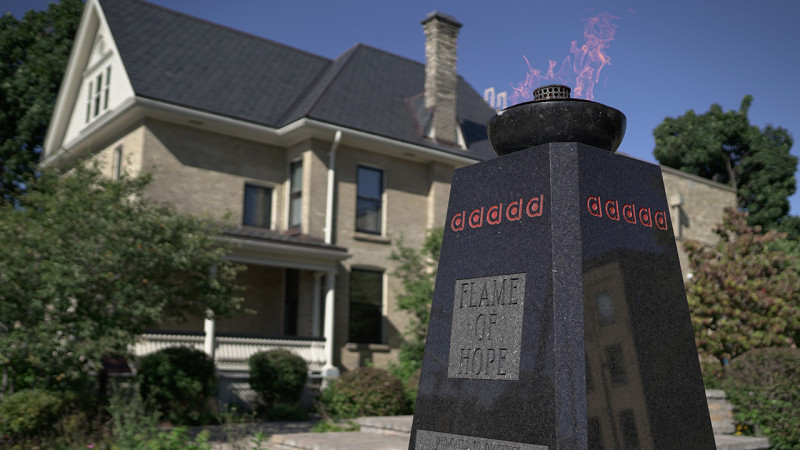 A dark grey marble monument with an engraved sign saying, "Flame of Hope" and a flame lit on the top stands in front of a older house surrounded by trees