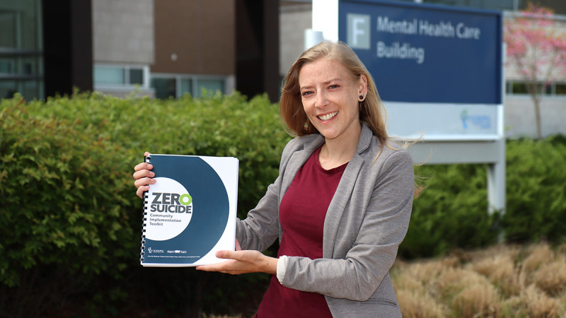 Shauna Graf holding a copy of a book called the Zero Suicide Implementation Kit, standing in front of the Mental Health Care Building at Parkwood Institute