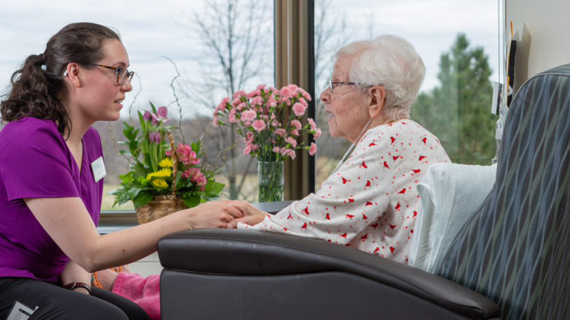 Young caregiver holding hands and talking with elderly woman seated in a chair, with flowers on the windowsill in the background.