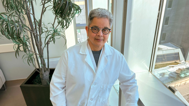 Savita Dhanvantari, PhD, in a white lab coat standing near a large window with a potted plant and a view of a building outside.