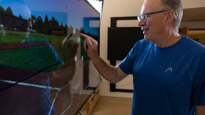 A person in a blue shirt pointing at a large screen displaying a sports field image, in a room with a blackboard and furniture.