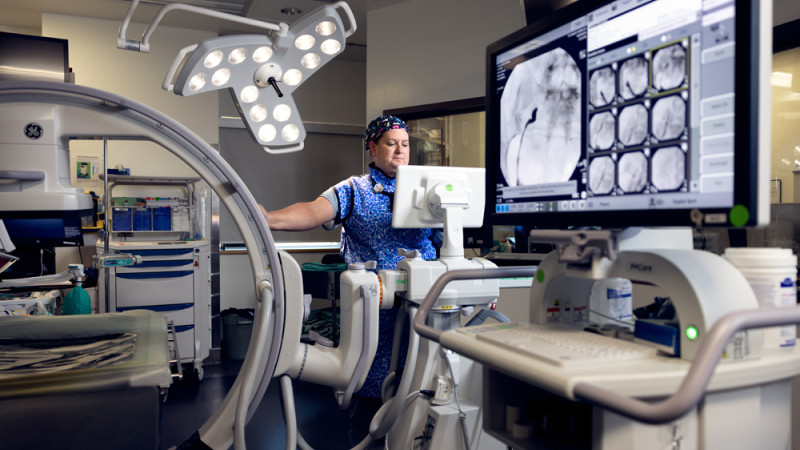 A medical professional in a hospital operating room, operating advanced imaging equipment with a large monitor displaying X-ray images.