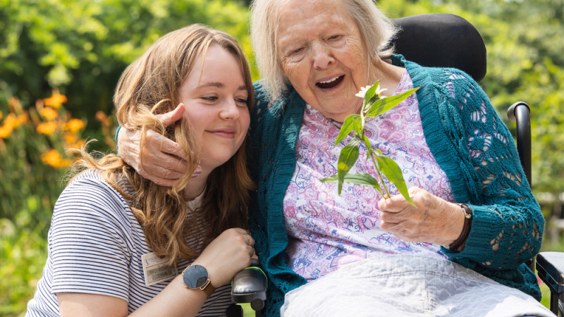 A young woman, wearing a striped shirt and name badge, sits next to an elderly woman in a wheelchair. The elderly woman is holding a small plant and smiling with delight.