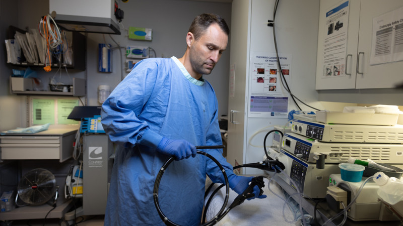 A medical professional in blue scrubs and gloves handling an endoscope in a clinical room with medical equipment and safety posters on the wall.