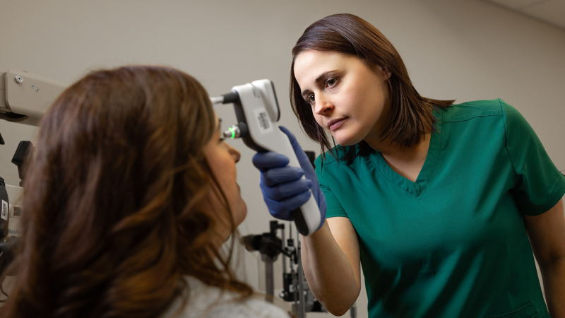 A medical professional in green scrubs uses a handheld tonometer to measure the eye pressure of a seated patient.