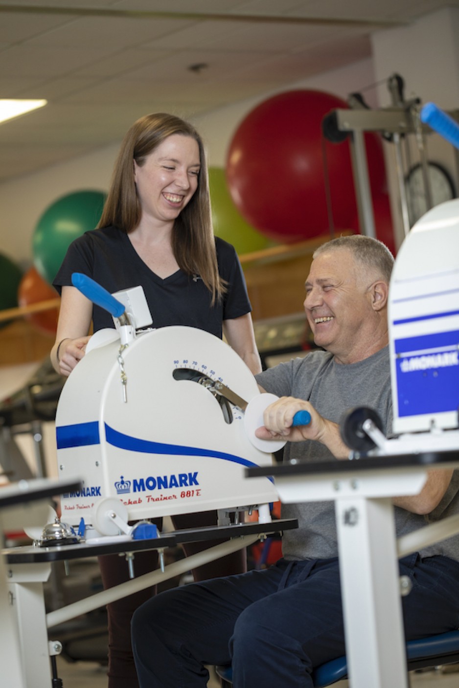 Man sitting by exercise equipment, biking with the arms. Woman standing next to him