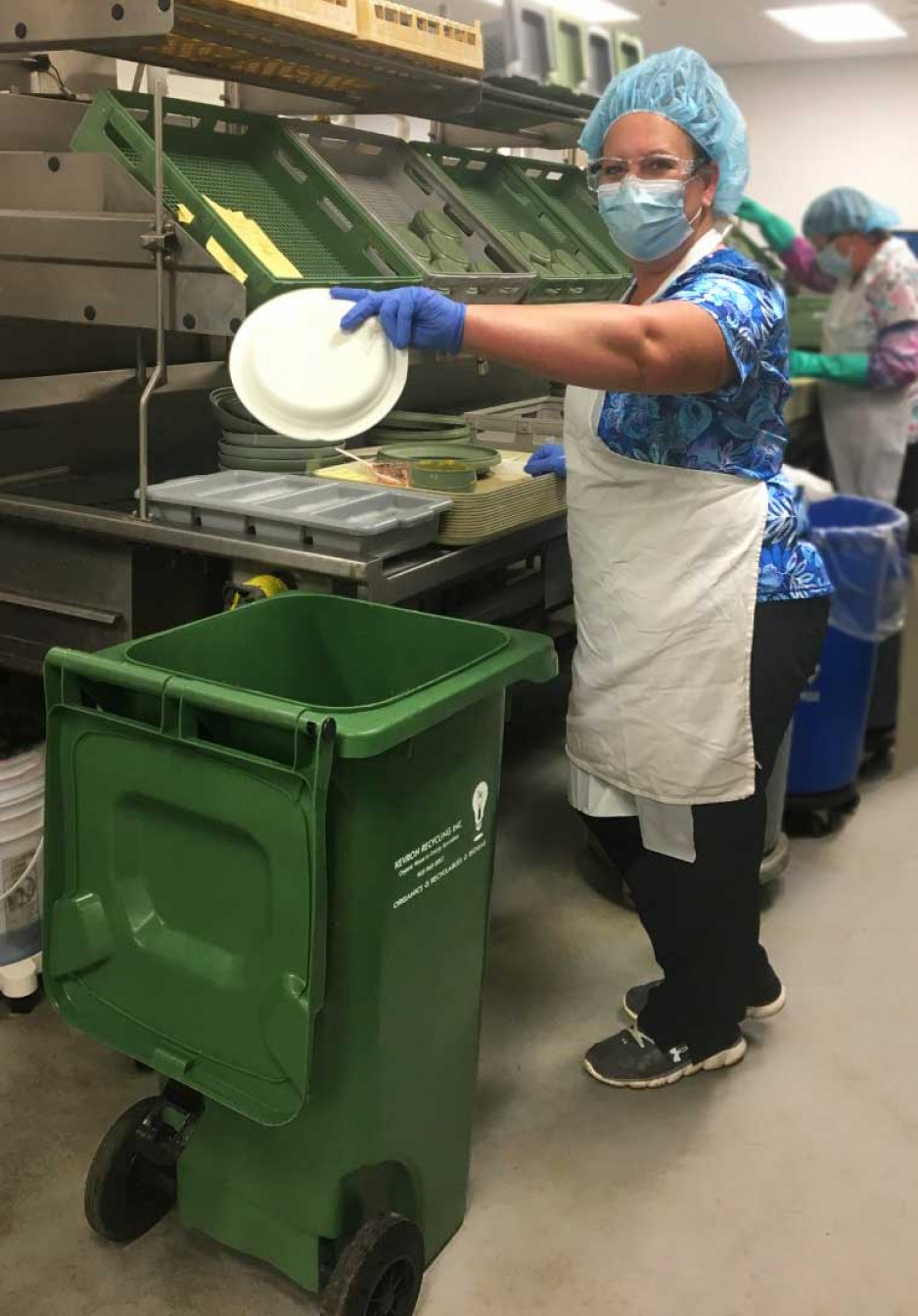 A food services worker in a hospital kitchen holding a plate over a green organics bin