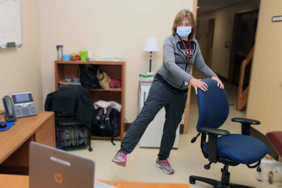 A physiotherapist demonstrates a leg raise exercise while looking at her patient on a laptop monitor