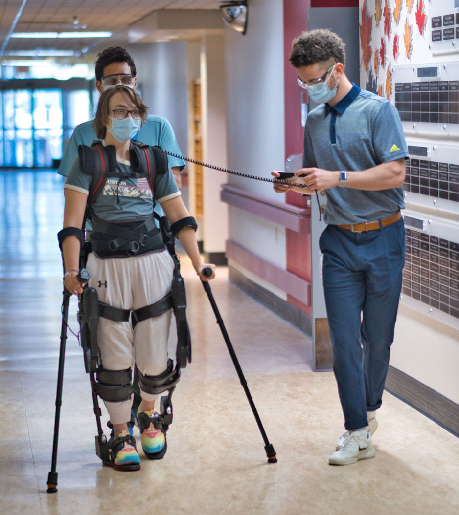 a female patient wearing an exoskeleton that allows her to walk, supported by a physiotherapist standing behind her, and another physiotherapist operating the controls