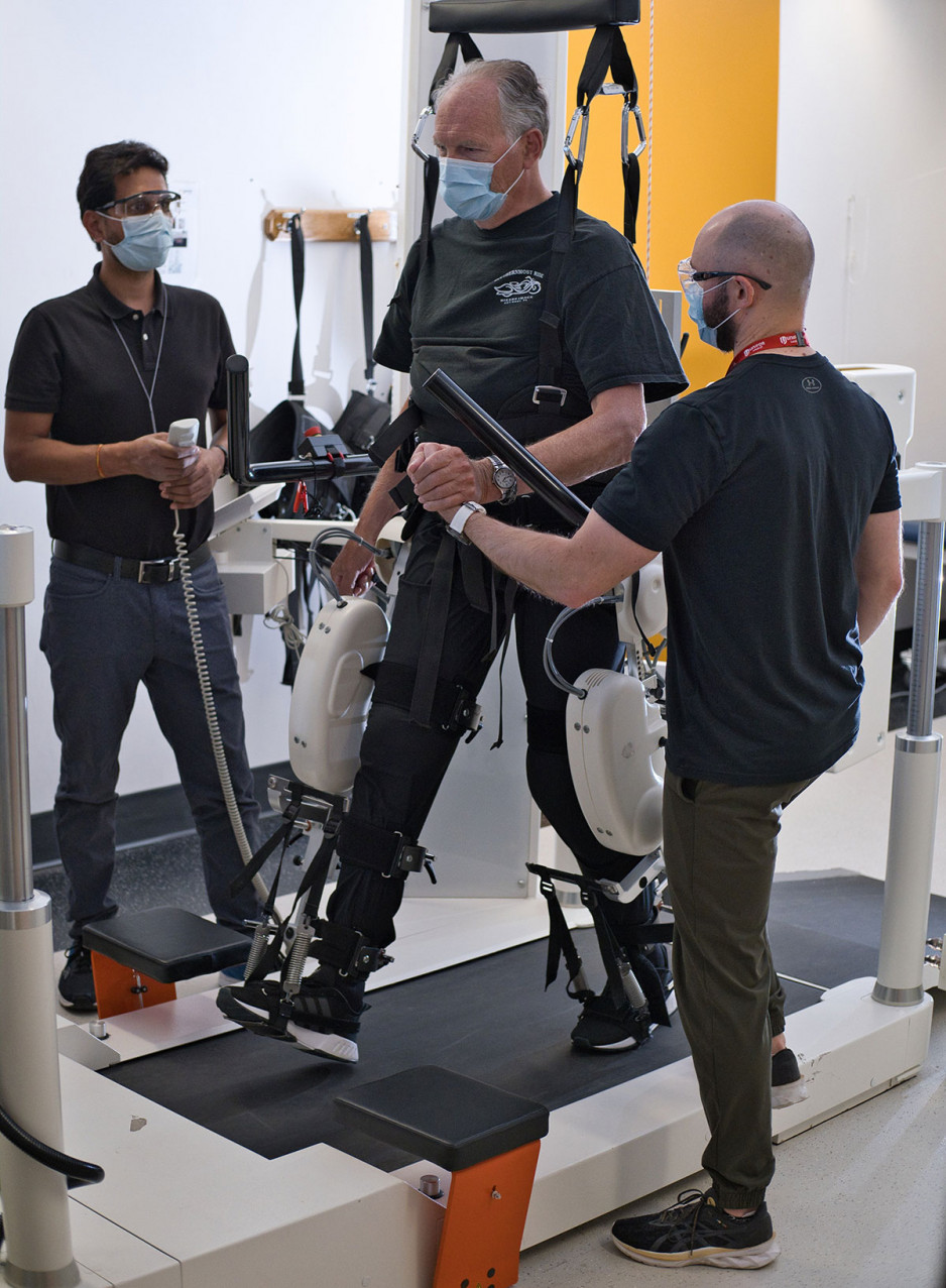 a male patient walking on a rehabilitative treadmill in a support harness, assisted by two physiotherapists