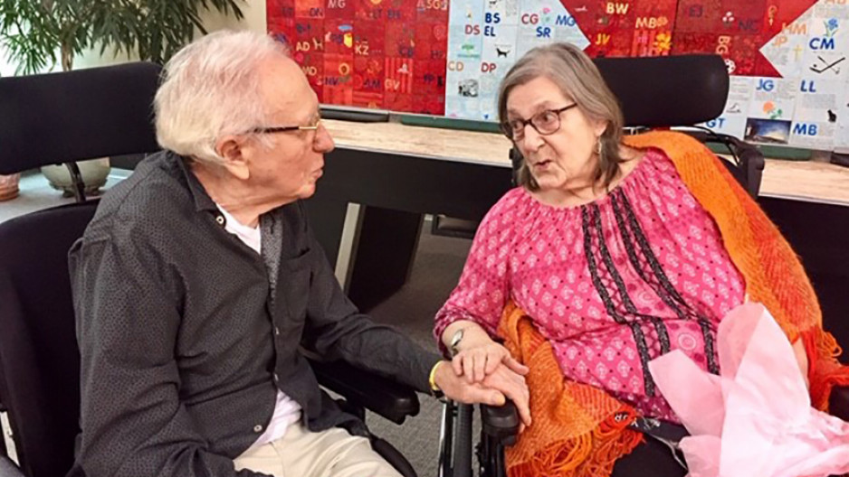 An elderly man and woman seated next to each other hold hands and gaze at each other. The elderly woman is facing the camera and the side profile of the man is visible.