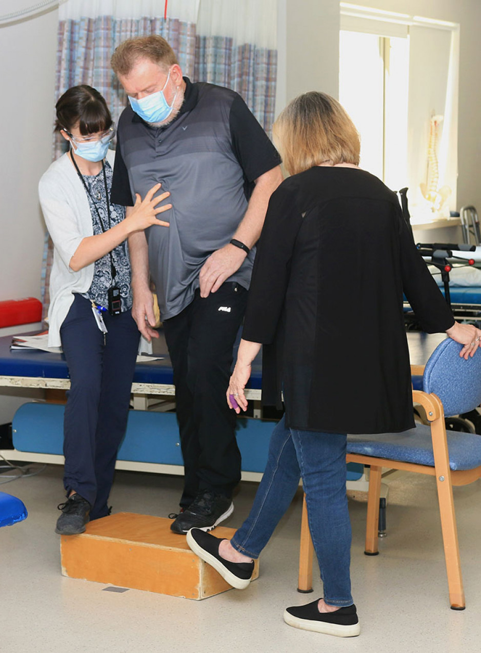 Paul Garrett using a step for physiotherapy, assisted by a therapist and his wife