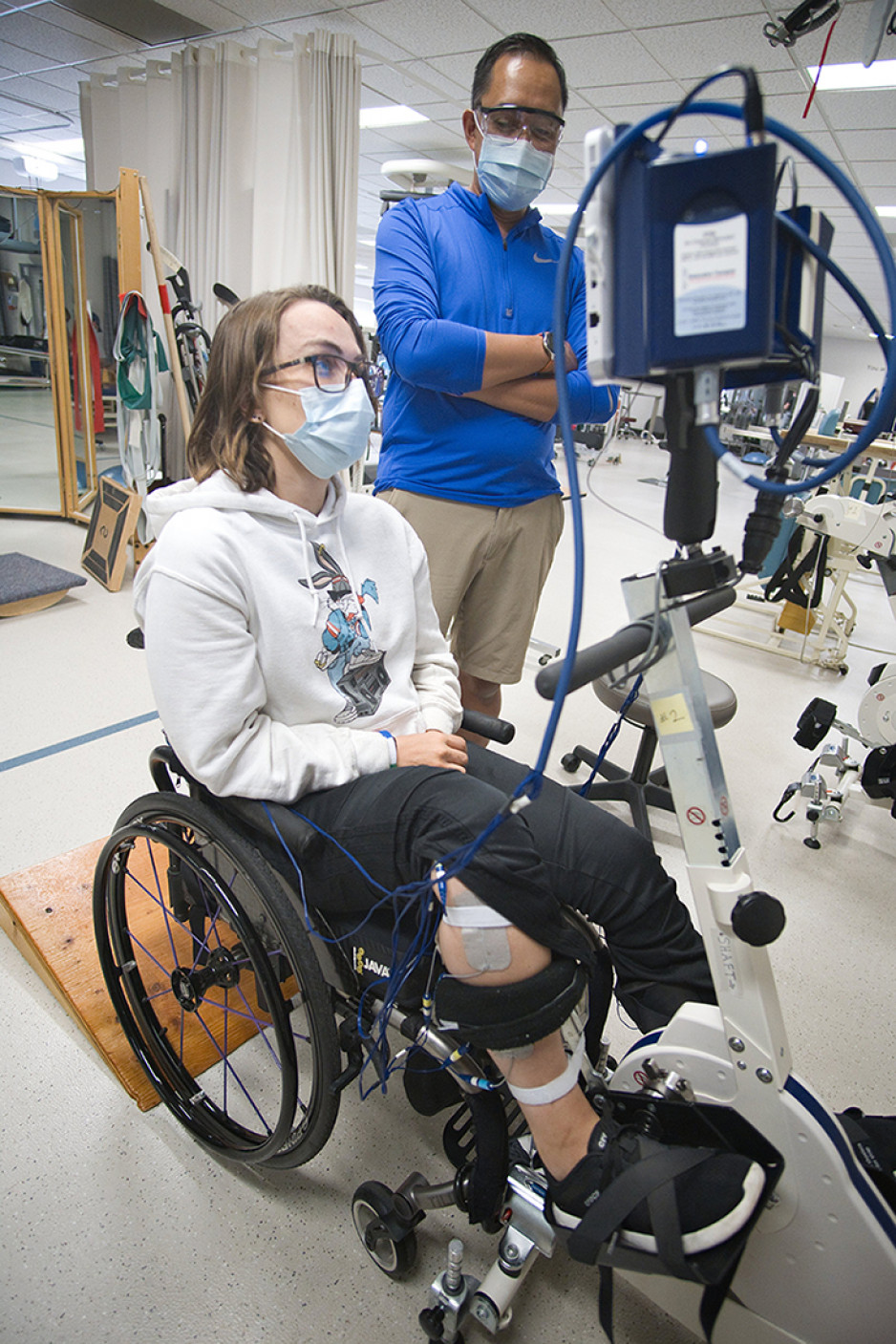 A masked woman in sweatpants and glasses in a wheelchair has her feet on the pedals of a modified exercise bike, which is connected to monitoring equipment. A masked man standing beside the equipment watches the woman