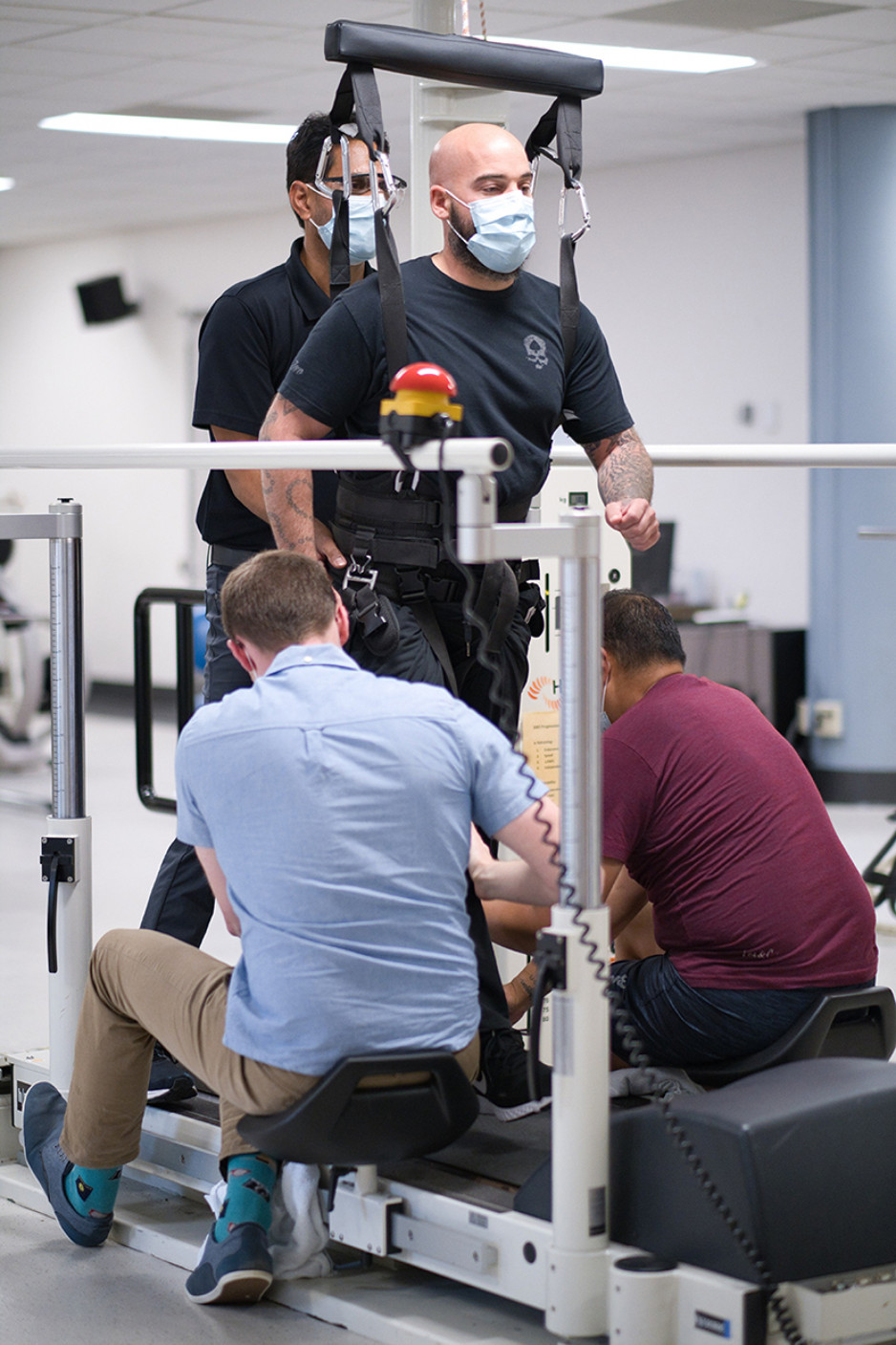 A masked man in a harness attached to the ceiling stands on a treadmill, as two men whose backs are to the camera crouch in front of him adjusting the device on him 