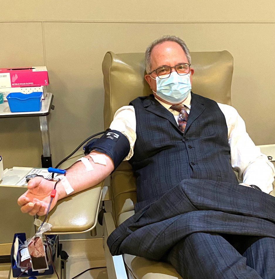 A  masked man with glasses in a suit vest and pants with a white dress shirt reclines on a hospital bed with his arm outstretched as he gives blood
