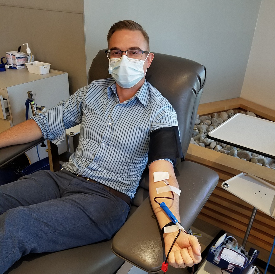 A masked man in a blue dress shirt and dress pants reclines on a hospital bed with his arm outstretched donating blood