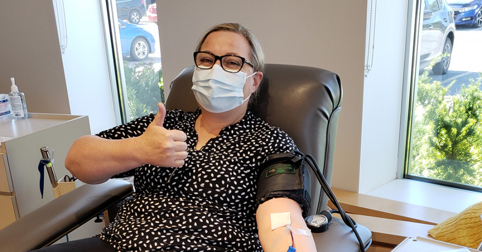  A masked woman in glasses, dress pants and a blouse reclines on a hospital bed with one arm outstretched as she gives blood and the other giving a thumbs up