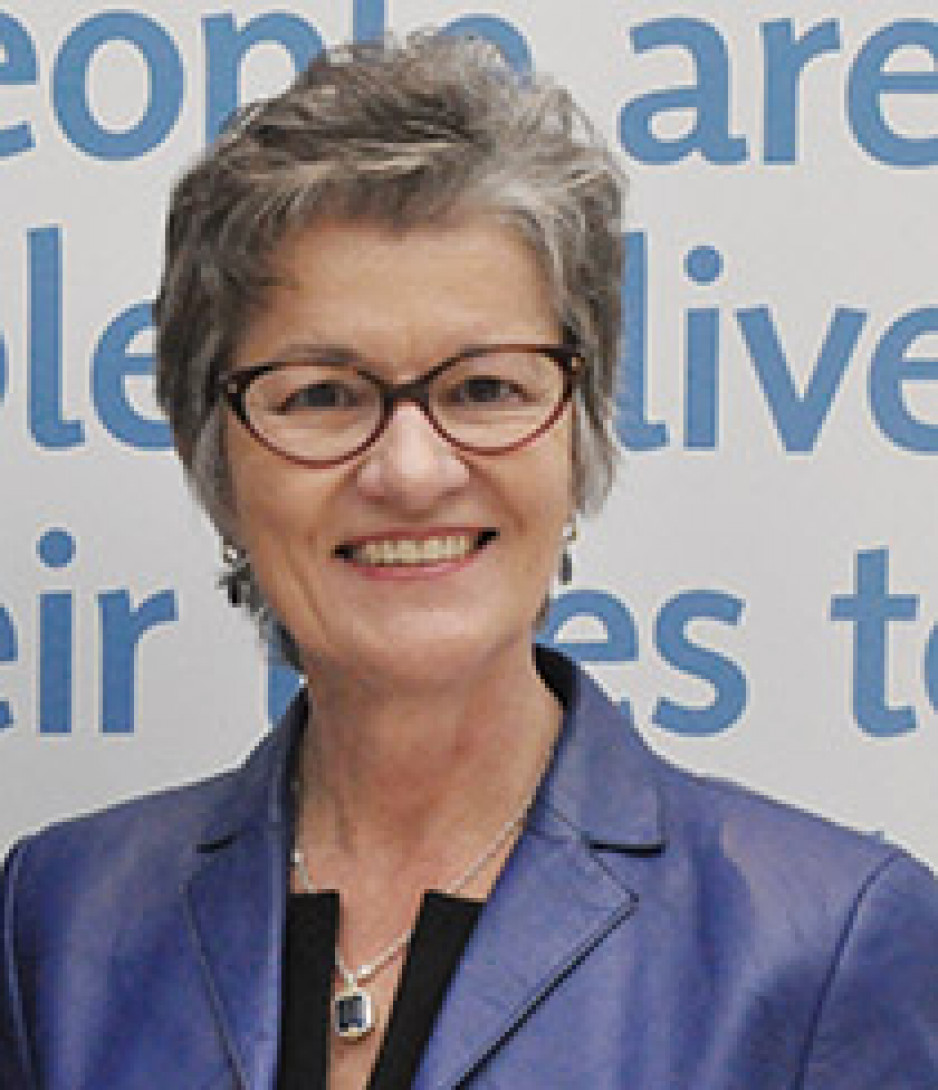A grey-haired woman wearing glasses and a blue shirt smiles at the camera