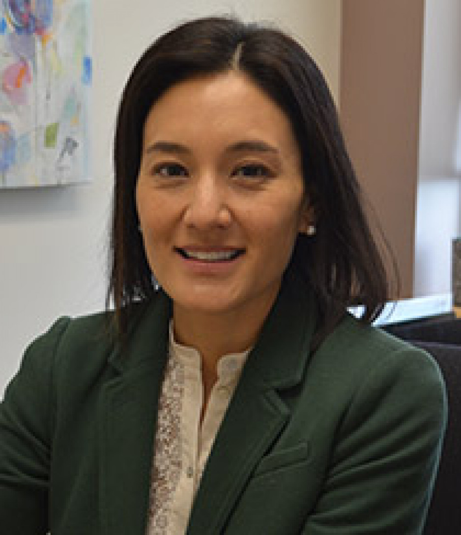 A woman with black hair in a dark blazer with a white shirt looks at the camera
