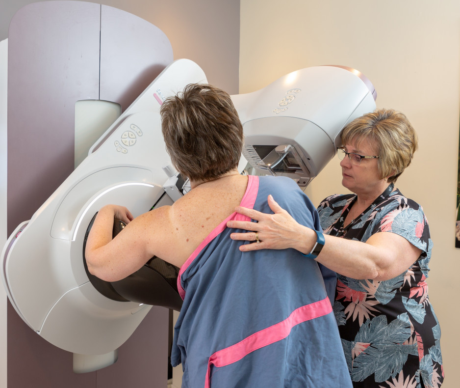 a woman wearing a shoulderless hospital gown stands before mammogram equipment with another woman in scrubs standing beside her and with one hand on the woman's back