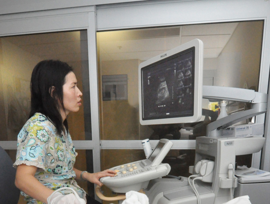 A woman with medium length black hair dressed in medical scrubs looks at a large monitor attached to equipment