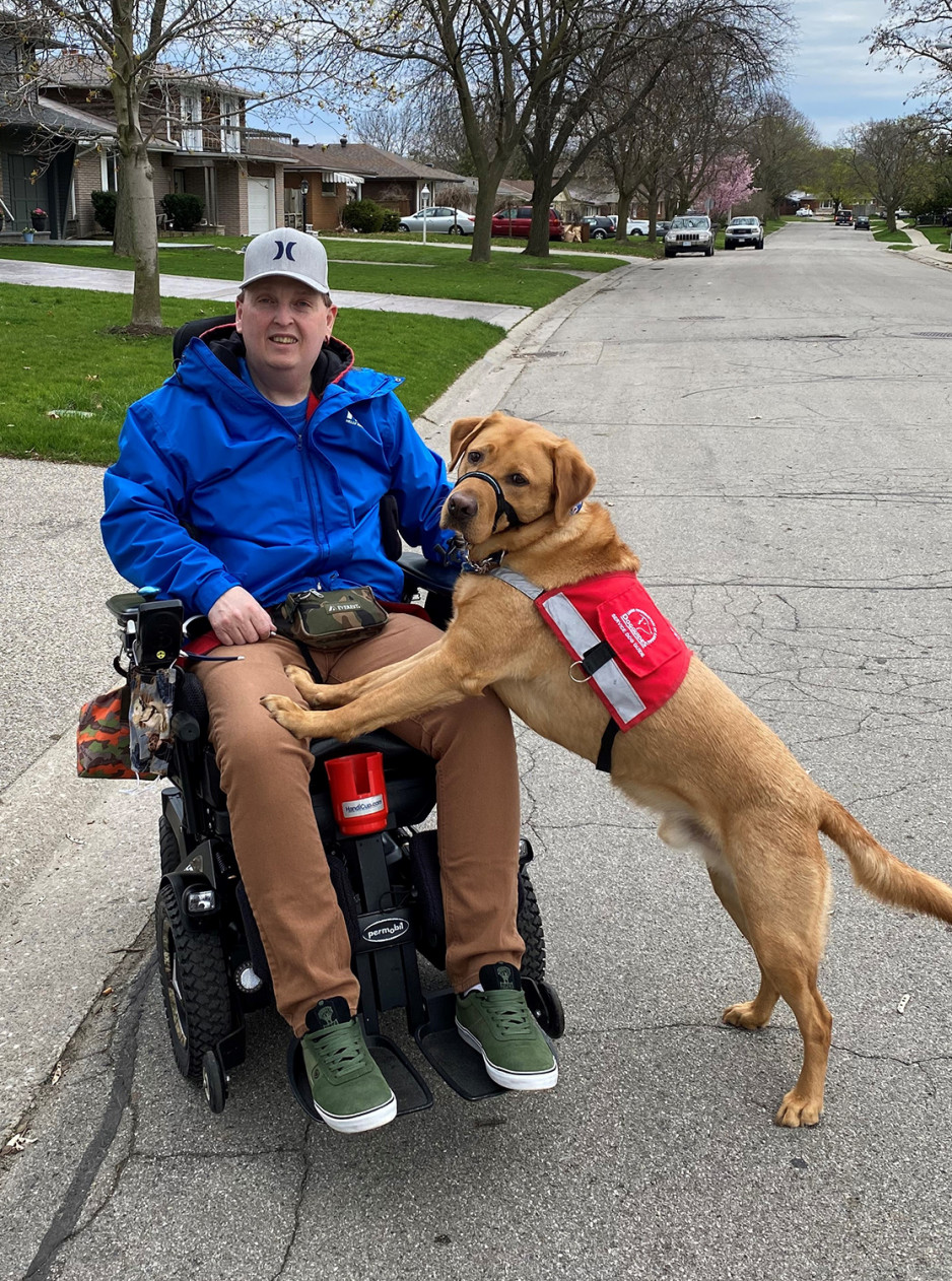 Phil Raney sitting in a wheelchair with his service dog beside him with his front paws on Phil's lap