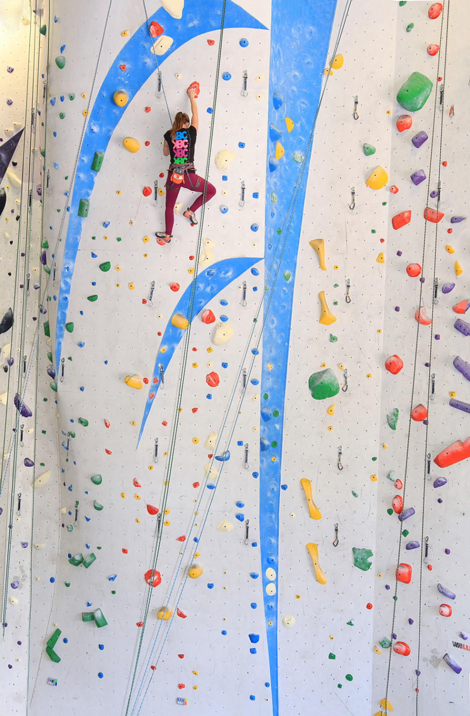 wide view of a high climbing wall with Miranda nearly at the top of the wall