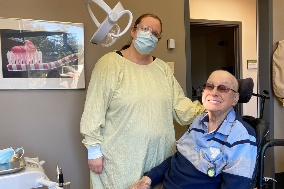 a veteran sitting in a wheelchair in the dental clinic, smiling with the dental light highlighting his teeth, and the dentist wearing a gown and mask standing beside him