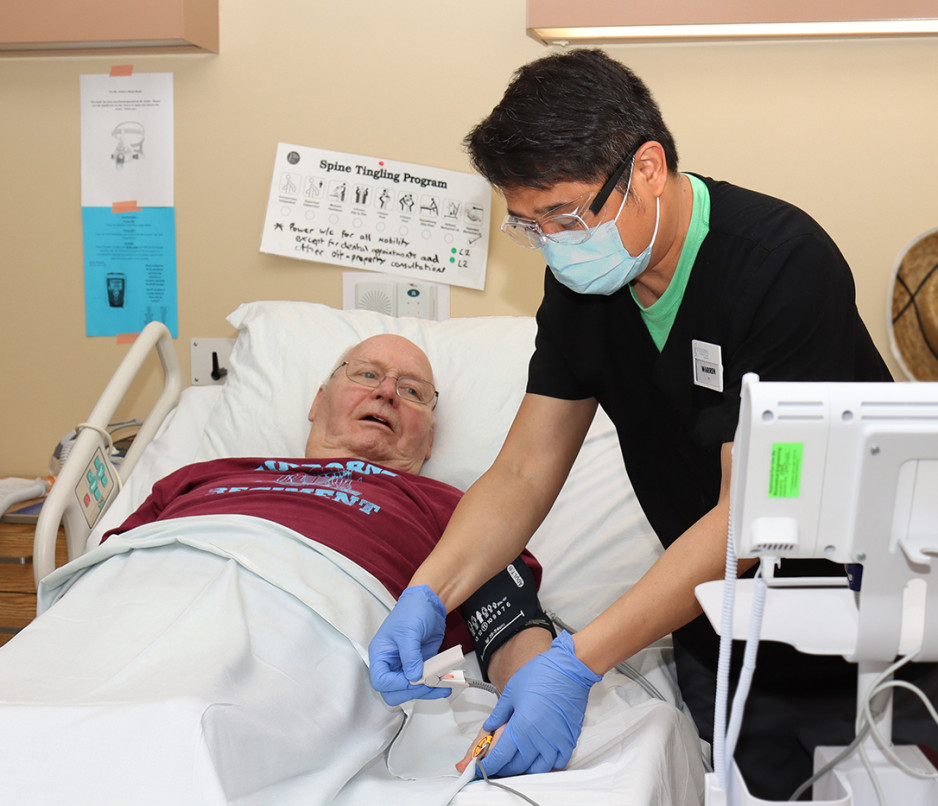 a male nursing taking the blood pressure of an elderly man sitting partly upright in a hospital bed in the Veterans Care Program