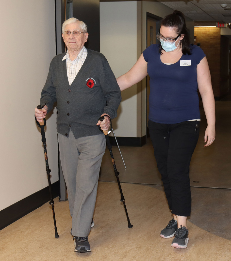 a veteran using two walking sticks walking down a hallway accompanied by a physiotherapist