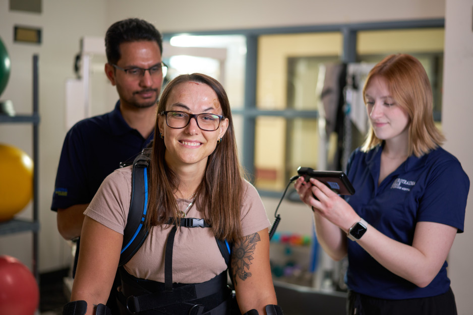 A woman smiles as she dons the exoskeleton supported by two physiotherapists