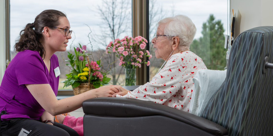 Young caregiver holding hands and talking with elderly woman seated in a chair, with flowers on the windowsill in the background.