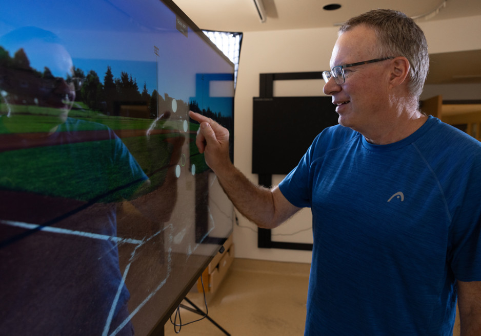 A person in a blue shirt pointing at a large screen displaying a sports field image, in a room with a blackboard and furniture.