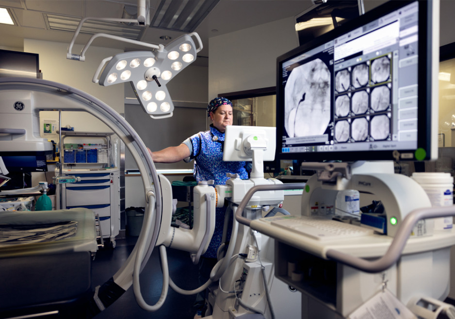 A medical professional in a hospital operating room, operating advanced imaging equipment with a large monitor displaying X-ray images.