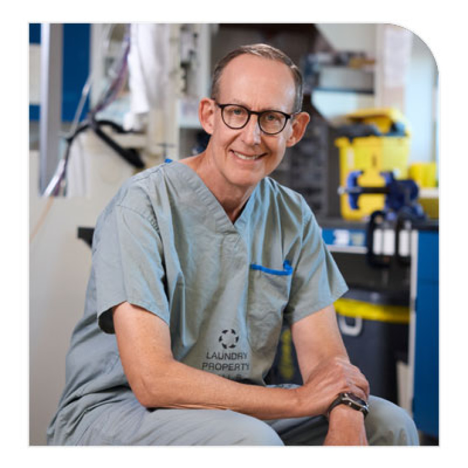 A smiling medical professional in scrubs with a "Laundry Property" label, sitting with arms crossed in a hospital or clinic setting.