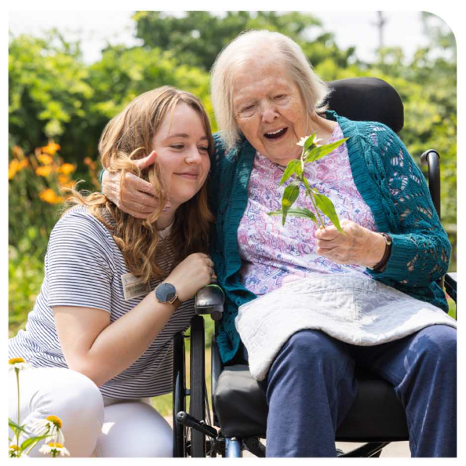 A young woman hugging an elderly woman in a wheelchair, both smiling, with the elderly woman holding a small plant in a garden setting.