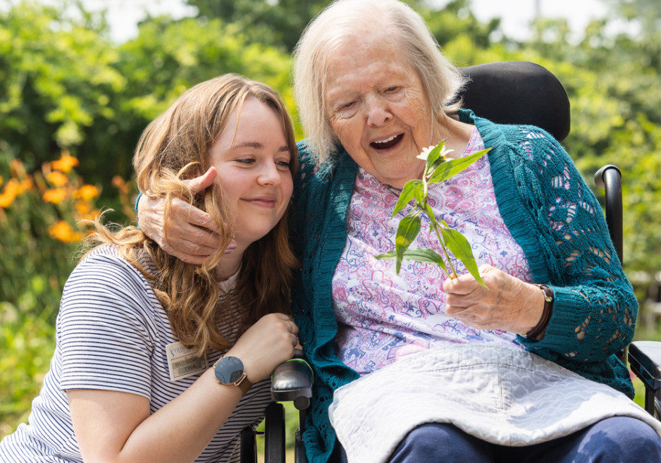 A young woman, wearing a striped shirt and name badge, sits next to an elderly woman in a wheelchair. The elderly woman is holding a small plant and smiling with delight.
