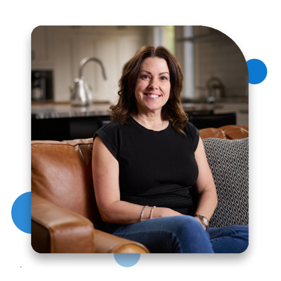 Krista Wilde sitting on a tan leather couch in a modern kitchen setting, wearing a black top and blue jeans, with a patterned cushion beside them.