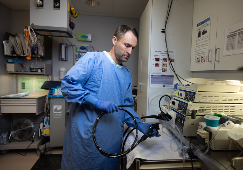 A medical professional in blue scrubs and gloves handling an endoscope in a clinical room with medical equipment and safety posters on the wall.
