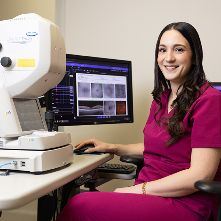A woman in maroon scrubs smiles while sitting at a computer next to a DRI OCT Triton medical imaging device.
