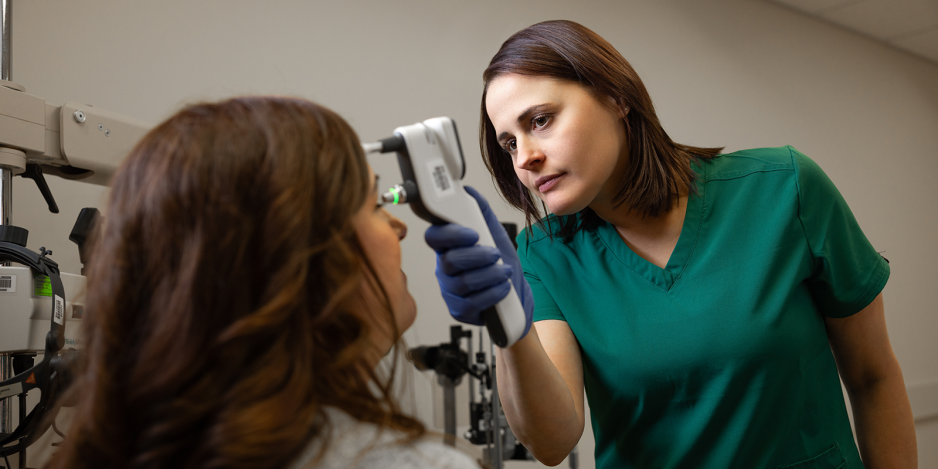 A medical professional in green scrubs uses a handheld tonometer to measure the eye pressure of a seated patient.