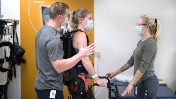a patient wearing a mobility exoskeleton, holding a walker, supported by a physiotherapist standing behind her and another physiotherapist in front of her