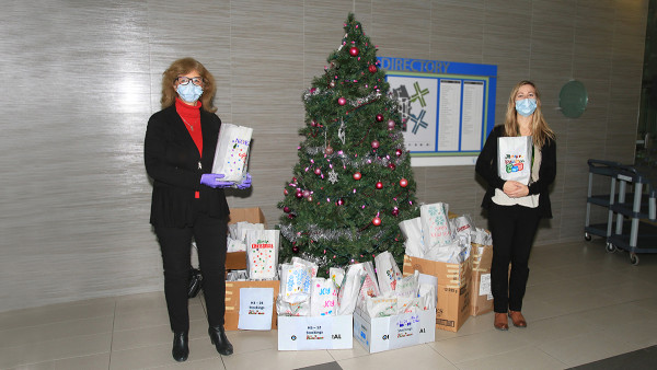 two women standing on either side of a Christmas tree holding gifts, surrounded by gift bags