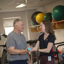 A man standing lifting weight with one arm, receiving support by another person